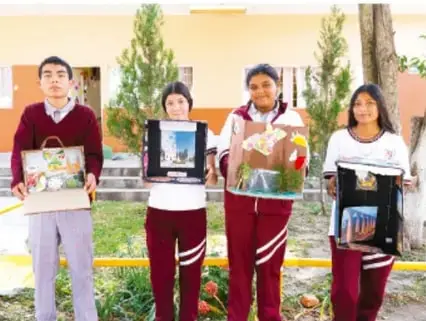 Fotografía de cuatro estudiantes de telesecundaria uniformados sosteniendo sus carteles/proyectos terminados frente a su escuela.