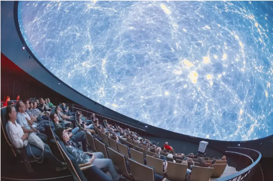 Fotografía del interior de un planetario: público sentado en butacas reclinadas mirando una cúpula enorme proyectada con un mapa estrellado azul brillante.