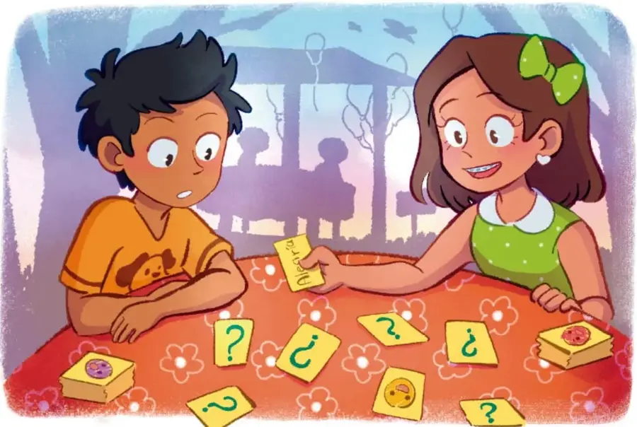 Niño con camiseta naranja y niña con moño verde juegan sentados a una mesa con mantel rojo floral: ella le entrega una tarjeta y sobre la mesa hay tarjetas amarillas con signos de interrogación, sugerencia del juego 'Adivina mi emoción'. Crédito: Karla Enríquez / Raúl Medina / TAMALLIPS.
