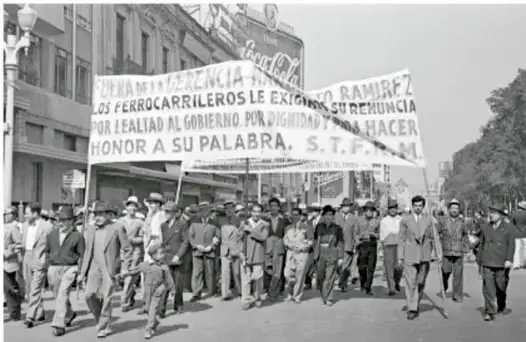 Fotografía en blanco y negro de una manifestación de ferrocarrileros. Los trabajadores marchan portando mantas con consignas como 'LOS FERROCARRILEROS LE EXIGEN SU RENUNCIA POR LEALTAD AL GOBIERNO POR DIGNIDAD Y HACER HONOR A SU PALABRA. S.T.F.R.M.'