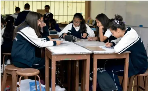 Estudiantes de primaria sentados en equipo alrededor de mesas, dialogando y tomando notas en el salón de clases.