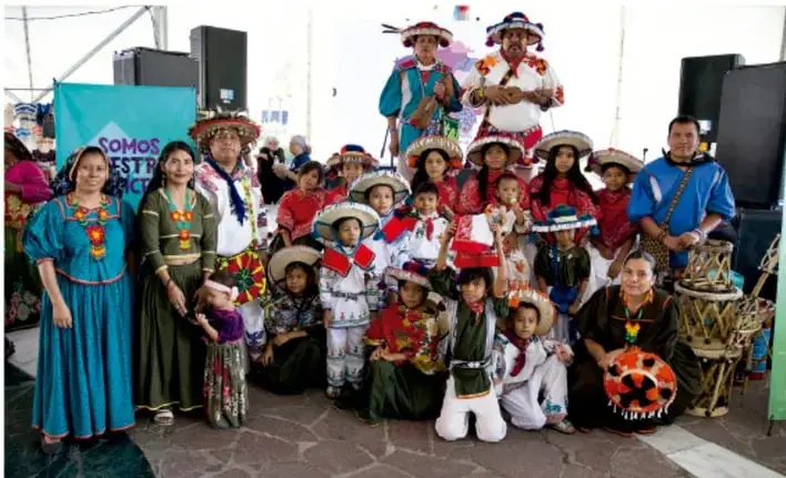 Fotografía de un grupo de niños, niñas y adultos con trajes típicos mexicanos y sombreros, frente a un lienzo con la frase 'Somos nuestro México', representando la diversidad cultural del país.
