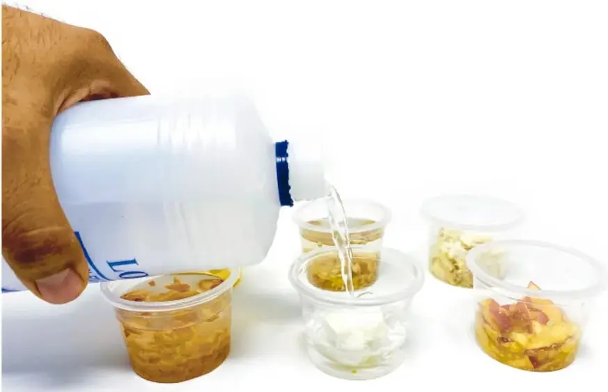 Mano vertiendo alcohol de un frasco blanco en vasos de plástico con alimentos machacados (pan, queso, fruta).