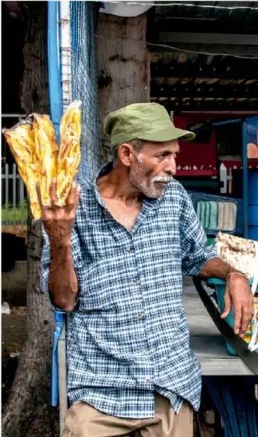 Fotografía de un hombre adulto de tez morena, con gorra verde y camisa a cuadros, que sostiene bolsas de producto (parece vender en la calle) junto a un puesto o vehículo, ilustrando a un afrodescendiente en su entorno cotidiano.