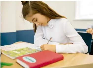 Niña escribiendo en un cuaderno sobre un escritorio, con libros y una tableta.