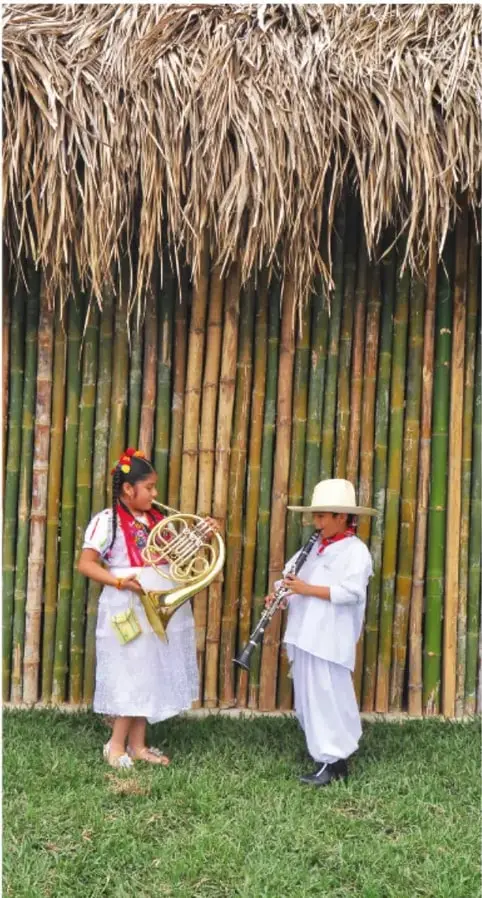 Dos niños con trajes tradicionales mexicanos tocando instrumentos musicales (trompa y clarinete) frente a una pared de carrizo con techo de paja.