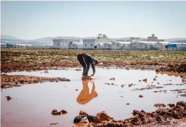 Fotografía de un paisaje con acumulación de agua de lluvia contaminada con residuos mineros, de color rojizo, y una persona agachada en el agua. Al fondo, edificaciones blancas y montañas.