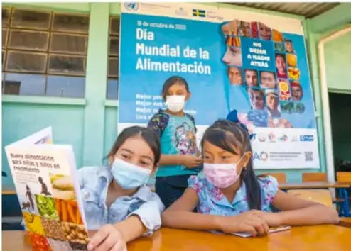 Fotografía de tres estudiantes con cubrebocas en un aula, frente a un cartel del Día Mundial de la Alimentación, sosteniendo un folleto sobre alimentación saludable.
