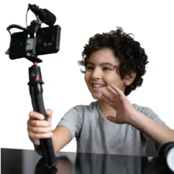 Niño con cabello rizado sostiene un palo selfie con una cámara y sonríe mientras graba.