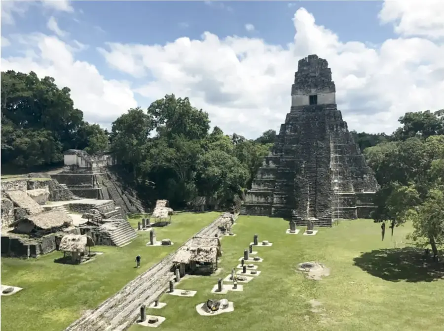 Fotografía de una zona arqueológica maya: una gran pirámide escalonada de piedra rodeada de árboles, con una plaza verde con estelas y estructuras menores, bajo un cielo con nubes.