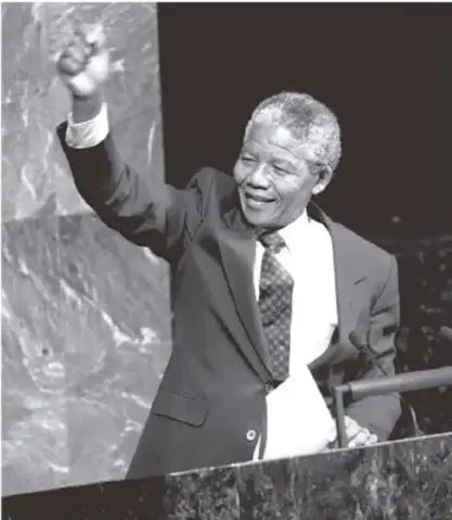 Fotografía en blanco y negro de un hombre sonriente de cabello blanco, con traje y corbata, que saluda con el puño en alto frente a un micrófono; por contexto se asocia a la lucha contra el apartheid (aparentemente Nelson Mandela).