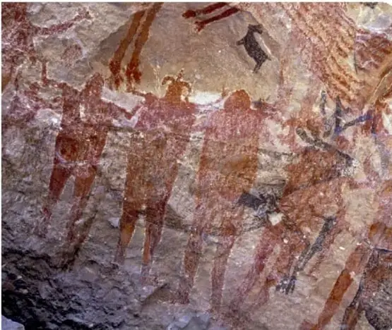 Pintura rupestre de la Cueva de las Monas, Chihuahua: figura humana central de color ocre con brazos extendidos, rodeada de líneas radiales y figuras humanas pequeñas, sobre roca clara.