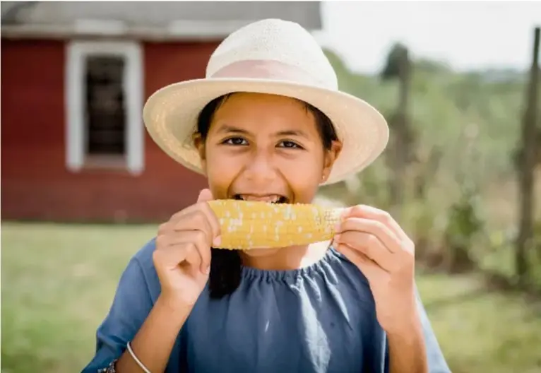 Fotografía de una niña con sombrero de paja comiendo un elote de maíz amarillo, en un entorno rural con una casa roja al fondo.