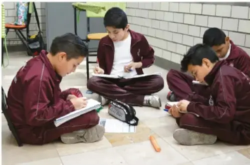 Fotografía de cuatro estudiantes con uniforme guinda sentados en el piso del patio escolar, escribiendo en sus cuadernos durante una actividad de observación.