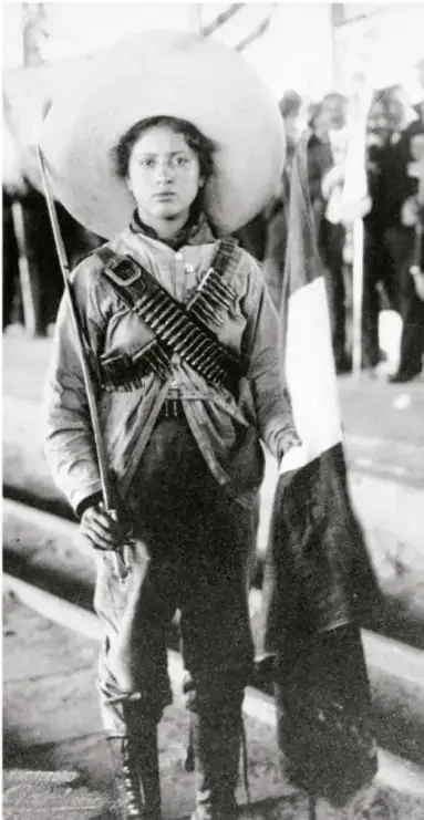 Fotografía en blanco y negro de una mujer revolucionaria con cananas cruzadas al pecho, sosteniendo un rifle y una bandera, con un sombrero grande; al fondo se ven otras personas.