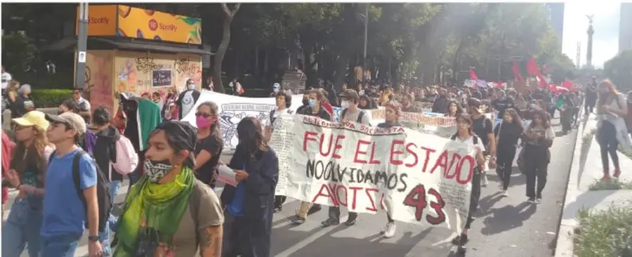 Fotografía de una marcha en la Ciudad de México. Personas caminan con pancartas; una de ellas dice 'FUE EL ESTADO' y '43 AYOTZINAPA'. Se observan edificios y árboles al fondo.