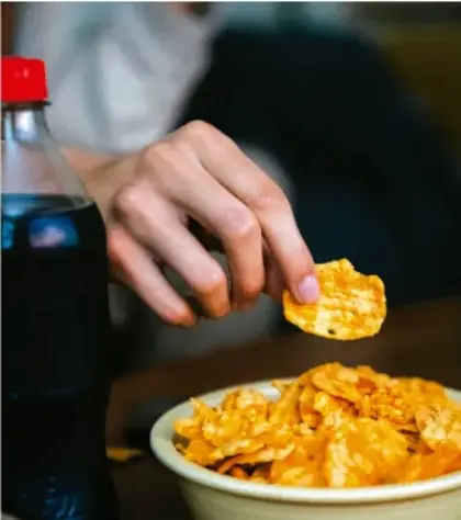 Fotografía de una mano tomando una botana de maíz de un tazón, con una botella de refresco al fondo, como ejemplo de alimentos ultraprocesados.