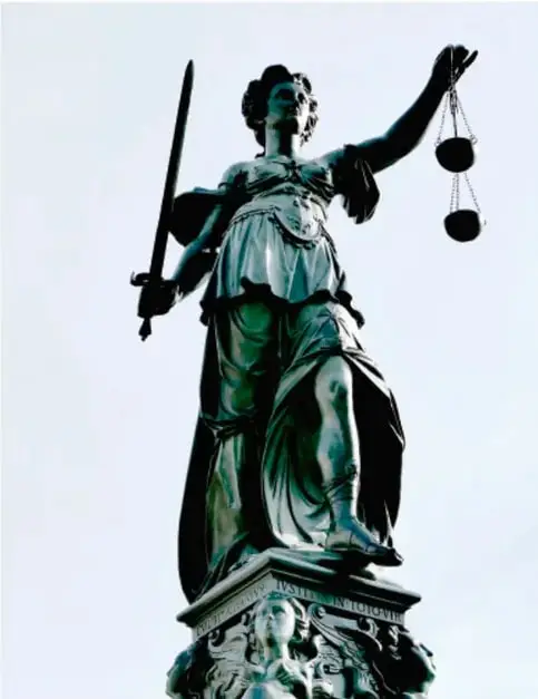 Fotografía de una estatua de la diosa de la justicia (Justitia) en tono verde bronce: sostiene una espada en la mano derecha y una balanza alzada en la izquierda, símbolo de imparcialidad y justicia.
