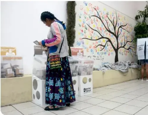 Mujer de espaldas con falda larga y blusa, frente a un puesto de revistas o libros en la calle. Hay un mural con un árbol y mensajes.