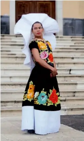 Fotografía de una mujer con vestido tradicional bordado y un tocado blanco, de pie sobre las gradas de un edificio: probablemente una performance o expresión cultural en vivo.