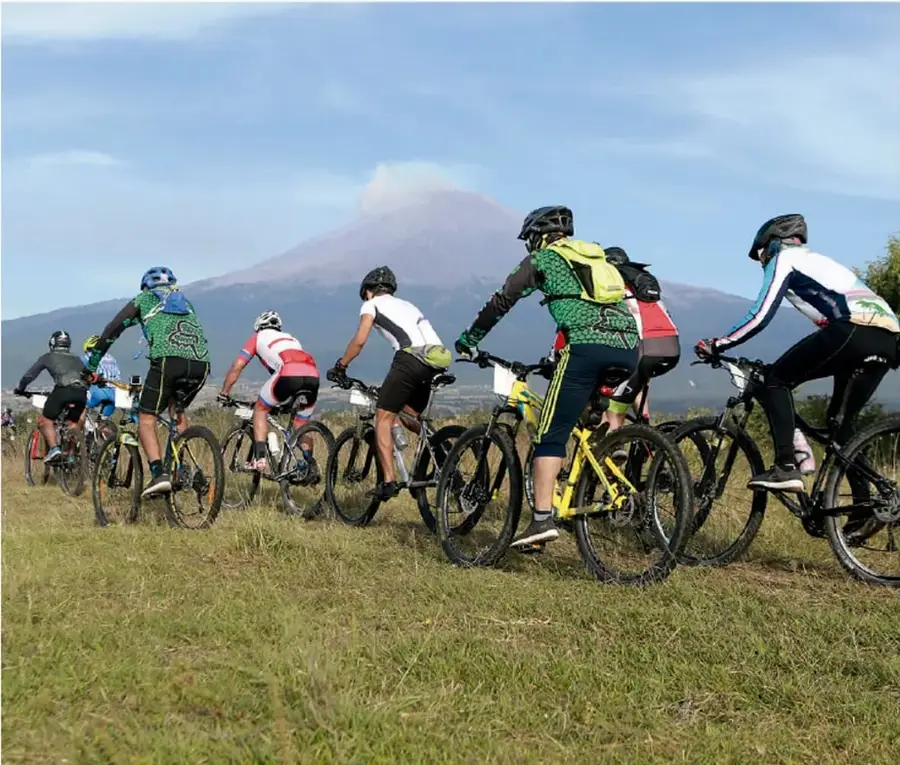 Fotografía de un grupo de ciclistas con casco y bicicletas de montaña, vistos de espaldas, sobre un terreno de pasto; al fondo se observa un volcán con una columna de vapor y un cielo azul con nubes.