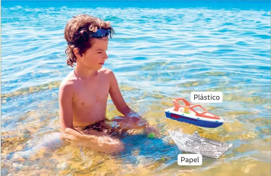 Fotografía de un niño con antiparras dentro del agua; junto a él flotan un barco de juguete de plástico (etiquetado 'Plástico') y un barco de papel de diario (etiquetado 'Papel').