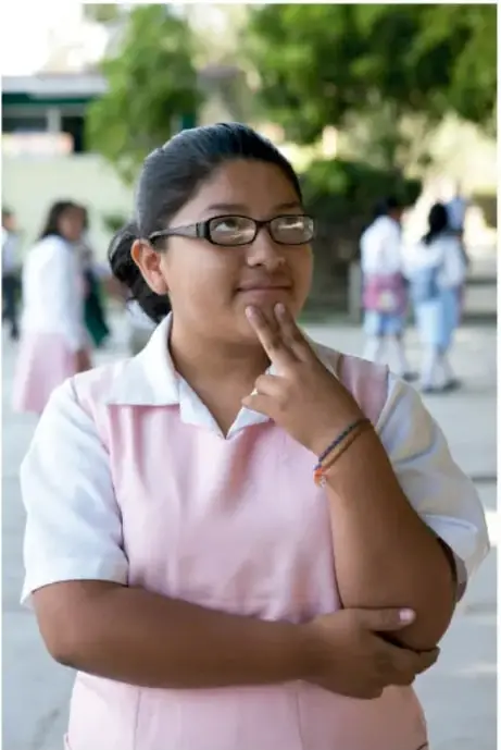 Fotografía de una estudiante de telesecundaria con lentes, en actitud pensativa, con el dedo en el mentón; estudiantes con uniforme al fondo en el patio escolar. Crédito: Francisco Manuel Palma Lagunes, Ciudad de México.