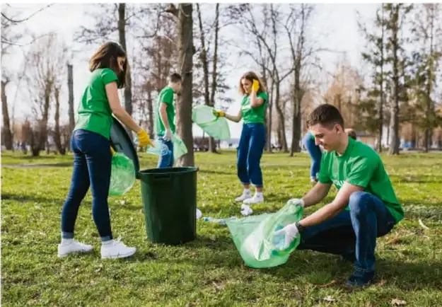 Grupo de jóvenes con camisetas verdes recogiendo basura en un parque, colocándola en bolsas y botes, ejemplo de ciudadanía ecológica en el ámbito público.