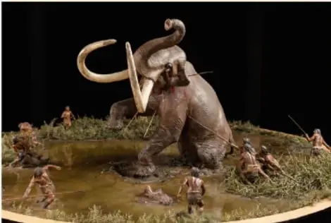 Recreación de un mamut atrapado en una trampa de barro, rodeado por cazadores con lanzas.