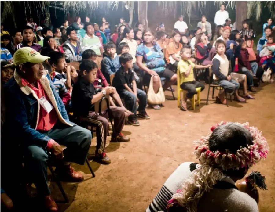 Fotografía de una asamblea comunitaria al aire libre: niñas, niños y adultos sentados en círculo en un terreno de tierra, escuchando a una persona con tocado tradicional que habla con micrófono.