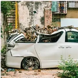 Auto blanco destruido junto a un muro en ruinas, ilustrando un choque inelástico.