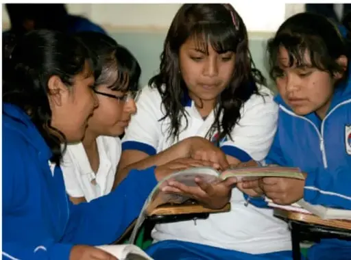 Fotografía de cuatro estudiantes (dos con uniforme azul) leyendo juntos un libro en el aula.