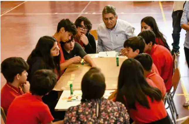Fotografía de un grupo de estudiantes de primaria sentados alrededor de una mesa en un patio escolar, dialogando con un adulto mayor que los guía durante una asamblea.