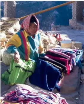 Fotografía de una artesana indígena sentada en un puesto de mercado, rodeada de textiles y prendas de colores, con una balanza de rueda al fondo.