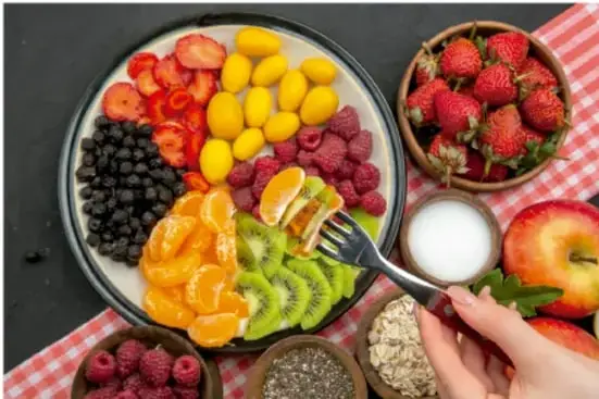 Fotografía de una mesa con un plato de frutas variadas (fresas, arándanos, kiwi, mandarina, uvas), un tazón de fresas, uno de leche y otro de avena; una mano sostiene un tenedor con un trozo de fruta.
