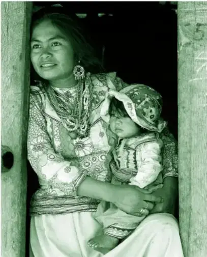 Fotografía (impresa en tinta verde): una mujer joven con blusa de flores, falda blanca, collares de cuentas y aretes grandes carga en brazos a un bebé con gorro estampado y trajecito de manta, enmarcados en la puerta de una casa.