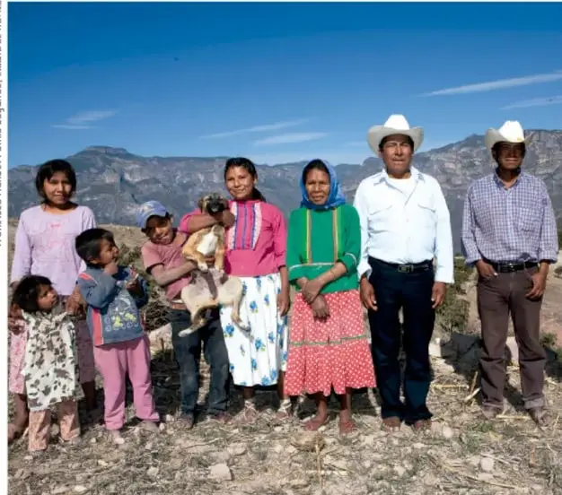 Fotografía de una familia y miembros de una comunidad rural (mujeres, hombres con sombrero, niños y un animal pequeño) de pie al aire libre con montañas y cielo azul al fondo. Crédito: Francisco Manuel Palma Luján, El País.