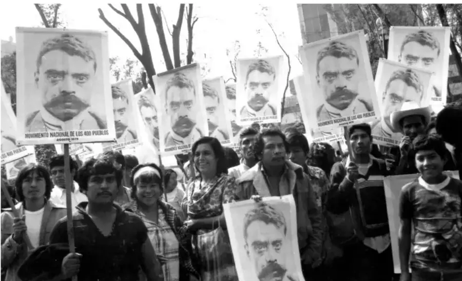 Fotografía en blanco y negro de una marcha con personas sosteniendo carteles con el rostro de un hombre y la leyenda 'Movimiento Nacional de los 400 Pueblos'.