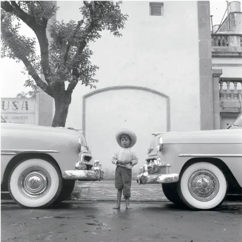 Fotografía en blanco y negro de un niño con sombrero de palma de pie entre dos automóviles antiguos de color claro. Detrás hay un árbol, una pared blanca con arco y un anuncio que dice 'USA'. Título: 'Entre el progreso y el desarrollo, ca. 1950, Héctor García (1923-2012)'.
