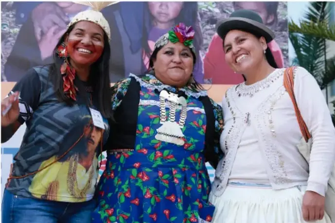 Tres mujeres con vestimenta tradicional indígena sonríen a la cámara; una de ellas lleva un tocado y otra un sombrero.