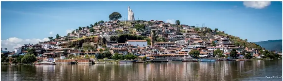 Fotografía panorámica de la Isla de Janitzio, Michoacán: cerro cubierto de casas coloridas que llegan hasta el agua del lago de Pátzcuaro, con la estatua blanca de la Virgen de la Salud en la cumbre, embarcaciones en la orilla y reflejo del paisaje en el agua; en la esquina inferior derecha de la foto aparece la firma del fotógrafo.