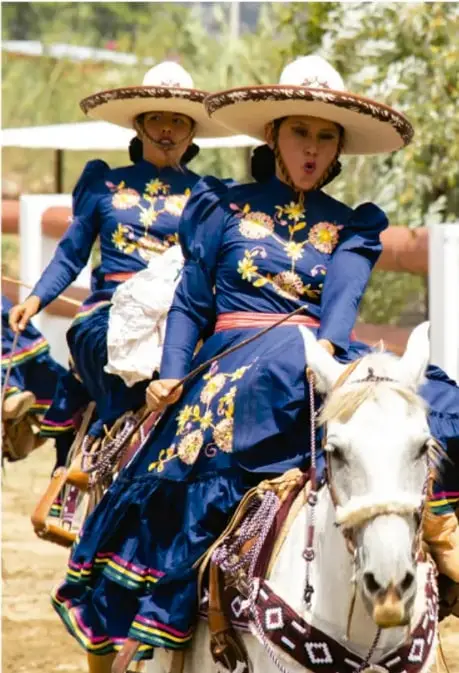 Dos personas vestidas con traje tradicional azul bordado y sombrero, montadas a caballo, ilustrando la cultura y las formas de vida de una comunidad.