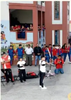Fotografía de estudiantes en el patio escolar participando en una actividad deportiva o presentación, con público observando desde un edificio.