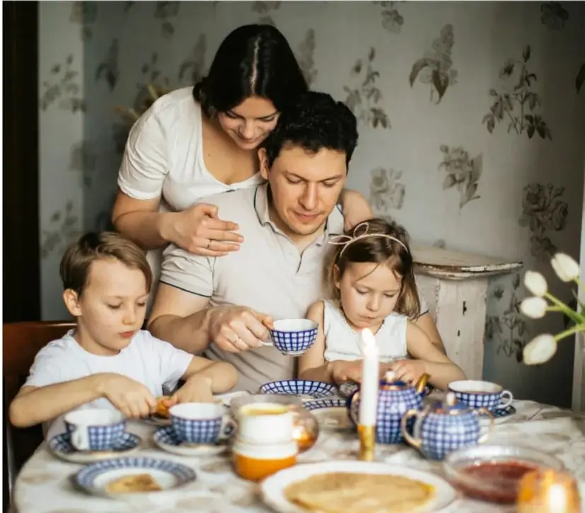 Fotografía de una familia desayunando junta alrededor de una mesa con tazas, pan y fruta.
