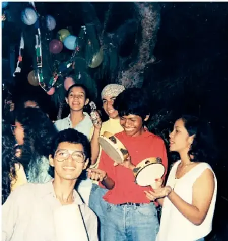 Fotografía de un grupo de jóvenes y niños tocando tambores y cantando en un evento comunitario nocturno, con globos y decoración.