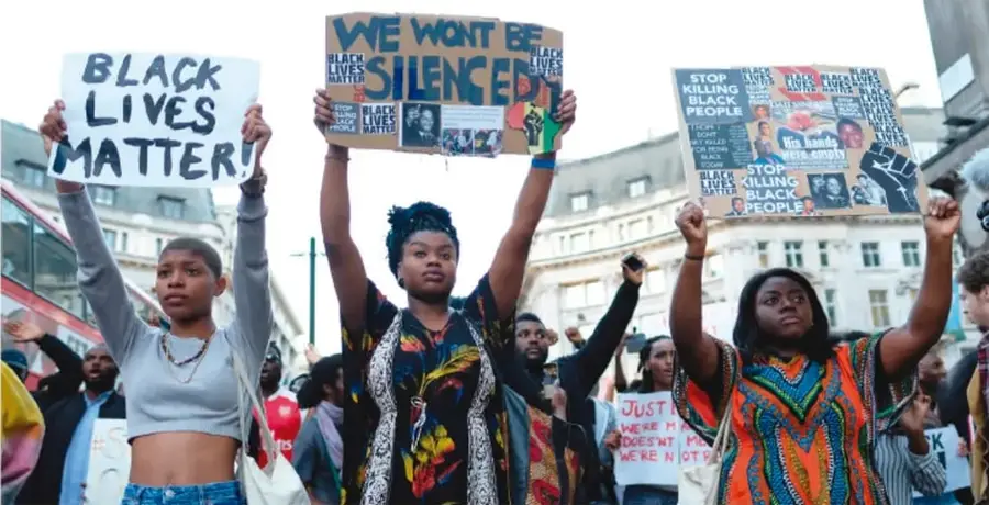 Fotografía de una protesta del movimiento Black Lives Matter: jóvenes afroamericanas alzan los puños y mantienen carteles con los lemas 'Black Lives Matter', 'We won't be silenced' y 'Stop killing black people', en una calle con edificios históricos.