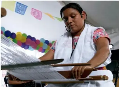 Fotografía de una joven artesana textil trabajando con un telar o bastidor, rodeada de colgantes y banderines de colores, representando a las portadoras del patrimonio cultural.