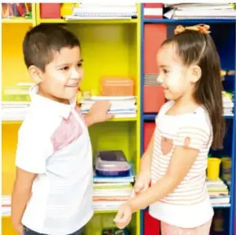 Fotografía de dos niños (niño con camisa blanca y niña con blusa rosa) conversando frente a un estante de libros de colores en el aula.