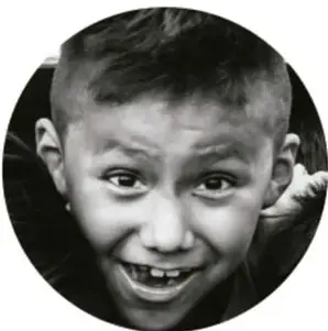 Fotografía en blanco y negro de un niño sonriente con las manos cerca de la cara, como si escuchara o hiciera sonidos.