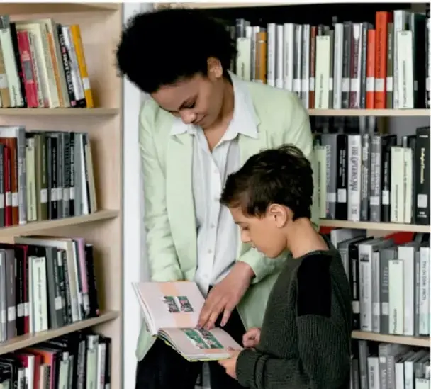 Fotografía de una mujer adulta y un niño de espaldas frente a un estante lleno de libros; la mujer sostiene un libro abierto y el niño observa. Representa la consulta en la biblioteca escolar.